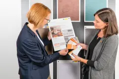 Zwei Mitarbeiterinnen von Sto Industrie stehen mit einem Farbtonfächer vor Musterplatten des Ausstellungssystems